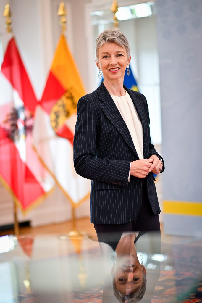 Frau mit kurzem grauen Haar und dunklem Nadelstreifen-Blazer steht in einem hellen Raum vor österreichischer, Kärntner und EU-Flagge, lächelnd in die Kamera.