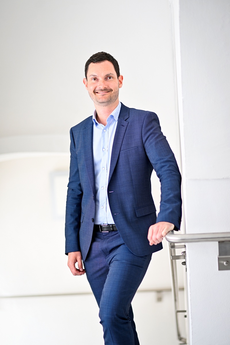 Stehendes Porträt: Mann im dunkelblauen Anzug und hellblauem Hemd, Hand am Metallgeländer, geht eine Treppe hinauf, lächelnd, neutraler heller Innenraum.